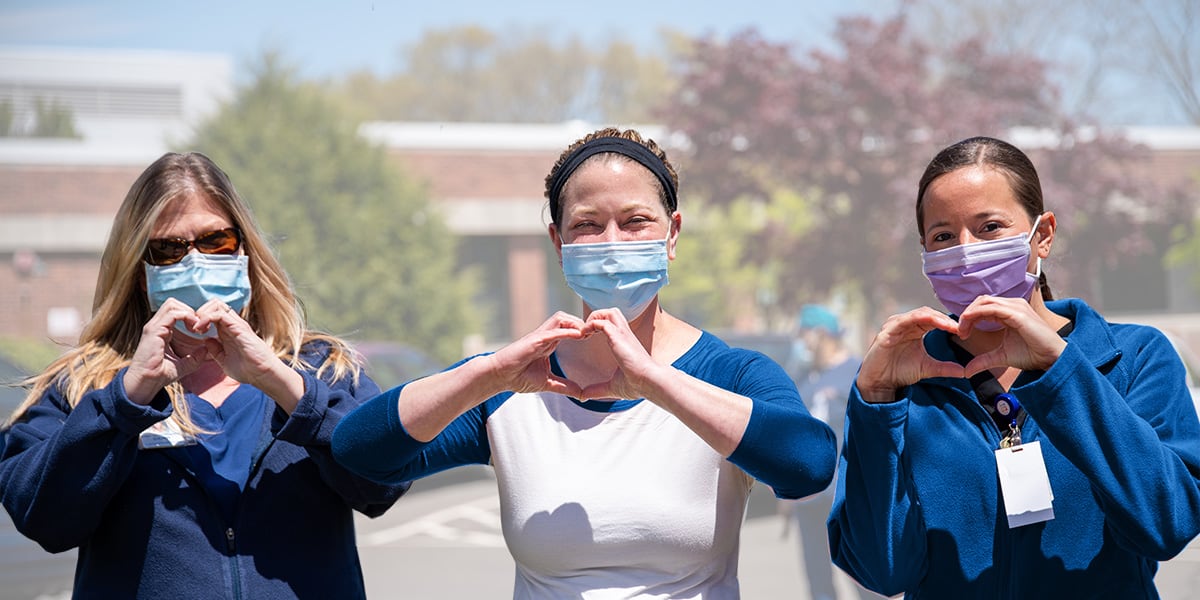 Three Personnel Support Workers wearing face masks and making the heart symbol with their hands.
